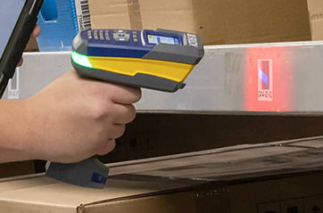 A person uses a Brady V4500 handheld barcode scanner to scan a label on a metal shelf. The scanner emits a red light onto the label, which contains a barcode.