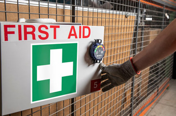 A person wearing a glove opens a first aid kit fastened to a manufacturing facility wall, representing part of an OSHA safety compliance plan.