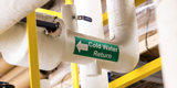 A green cold water return pipe marker on an overhead pipe in a manufacturing environment.